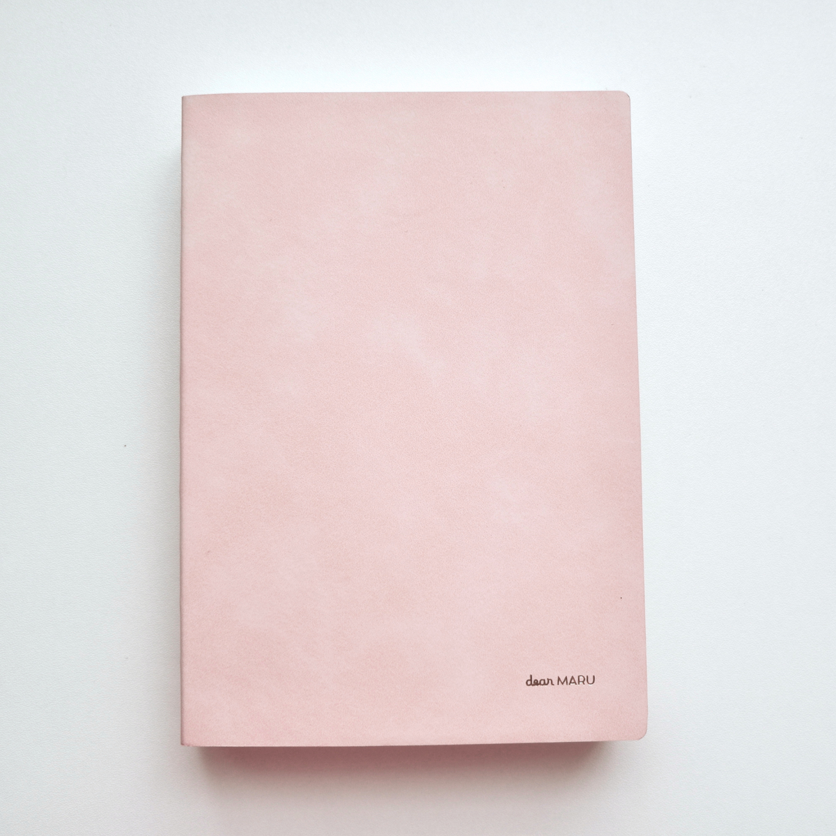 A notebook with a soft, buttery-textured pink leatherette cover with a small gold foil logo reading "Dear MARU" in the bottom right corner, photographed flat on a clean white background.
