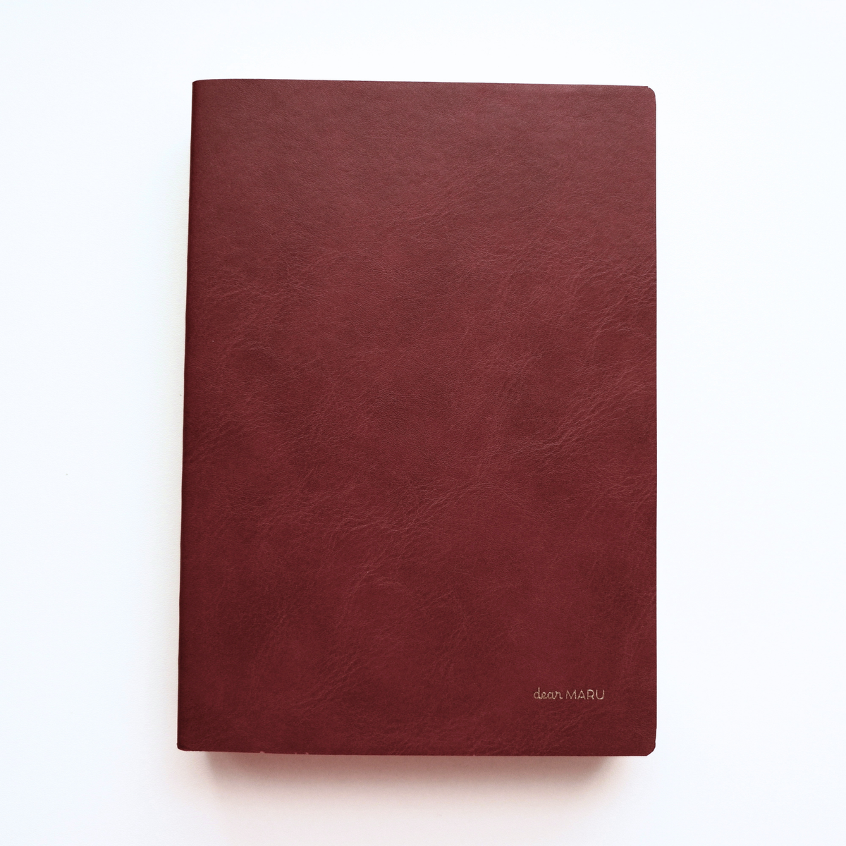A notebook with a soft, buttery-textured blood red leatherette cover with a small gold foil logo reading "Dear MARU" in the bottom right corner, photographed flat on a clean white background. 