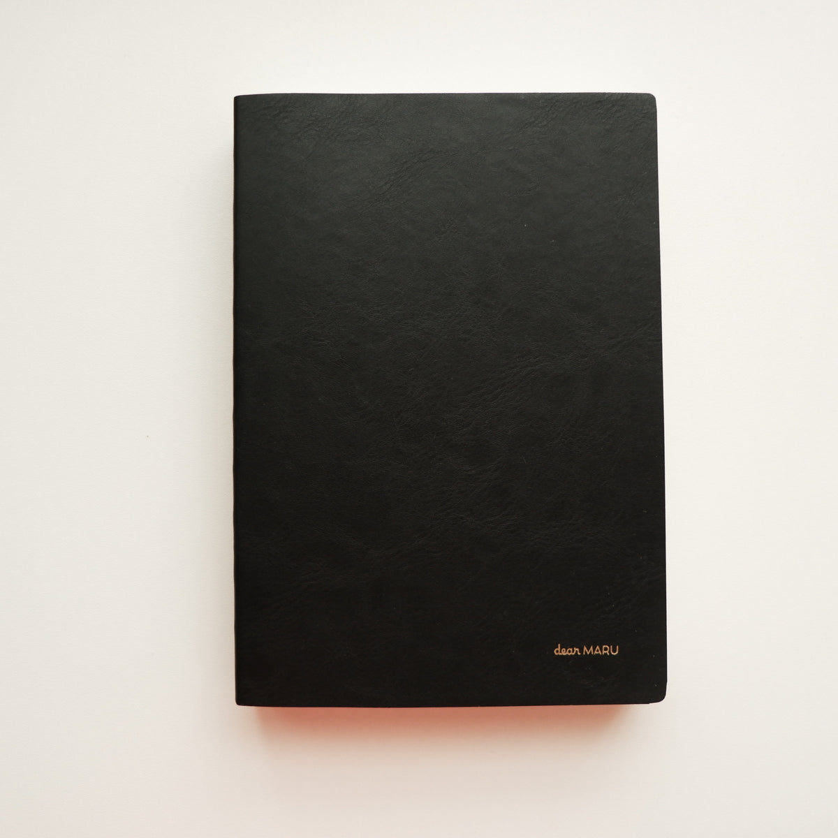 A notebook with a soft, buttery-textured black leatherette cover with a small gold foil logo reading "Dear MARU" in the bottom right corner, photographed flat on a clean white background. 